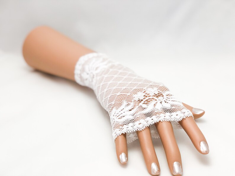 Puede incluir: Guantes sin dedos de encaje blanco con un dise&ntilde;o floral en el pu&ntilde;o y un patr&oacute;n de malla de diamantes. Los guantes se muestran en una mano de maniqu&iacute; con u&ntilde;as pintadas, sobre un fondo blanco.