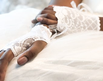 May include: Close-up of white lace fingerless gloves with delicate floral and geometric patterns. The gloves are tied with white ribbons and are resting on a white, textured surface, possibly a wedding dress.