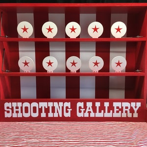 May include: A red wooden shooting gallery game with white stripes and eight white targets with red stars. The game has two shelves and the words "SHOOTING GALLERY" are printed in white letters on the front.