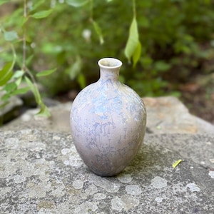 Könnte beinhalten: Eine kleine, dekorative Keramikvase mit rundem Körper und schmalem Hals. Die Vase hat eine hellgraue Grundfarbe mit einem strukturierten, kristallinen Muster in Blau- und Silbertönen. Die Vase steht auf einer Steinoberfläche.