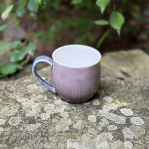 May include: A ceramic mug with a rounded body and a curved handle. The exterior features a muted mauve and lavender glaze with a subtle vertical texture, while the interior is finished in a smooth, clean white glaze with visible circular ridges.