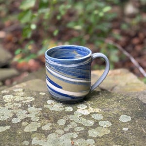 May include: A handmade ceramic mug with a blue, white, and olive green swirl pattern. The mug has a rounded body and a curved handle. The mug is sitting on a stone surface. The mug is approximately 4 inches tall.
