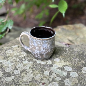 May include: A ceramic mug with a crystalline glaze featuring a white and grey starburst pattern on the exterior. The mug has a curved handle and a dark brown interior. The rim shows a thin band of light brown glaze, contrasting with the textured body.