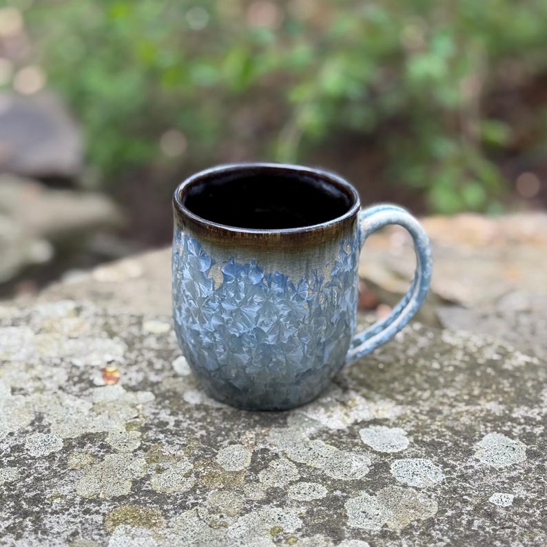 Puede incluir: Taza de cer&aacute;mica hecha a mano con un esmalte azul cristalino texturizado. El borde es marr&oacute;n oscuro y el asa es azul claro. La taza est&aacute; sobre una superficie de piedra.