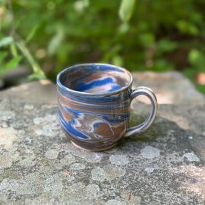 May include: A handmade ceramic mug with a marbled design. The mug features swirls of blue, brown, and gray. It has a rounded body and a curved handle. The mug is sitting on a stone surface, with a blurred green background.