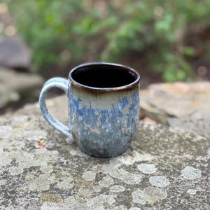 Puede incluir: Una taza de cer&aacute;mica con un esmalte cristalino azul y beige. La taza tiene un borde marr&oacute;n oscuro y un asa azul claro. La taza est&aacute; sobre una superficie de piedra con un fondo verde borroso.