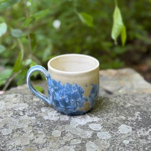May include: A handmade ceramic mug with a beige exterior and a blue crystalline glaze. The handle and the bottom of the mug are also blue. The mug is sitting on a gray stone surface, with green foliage in the background.
