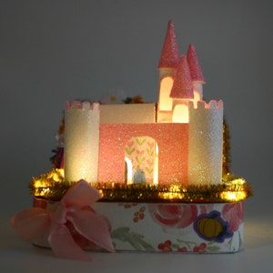 May include: A pink and white castle-shaped cake with a gold glitter trim and a pink bow. The castle has a white glitter roof and windows. The cake is decorated with flowers and a pumpkin.