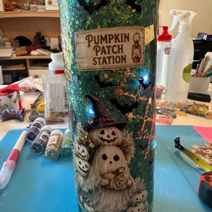 May include: A cylindrical tumbler with a green glitter finish featuring black bat silhouettes and a rectangular label that reads PUMPKIN PATCH STATION with a lantern icon. Below, a ghost wearing a witch hat holds a pumpkin labeled Boo.