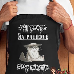 May include: Black apron with the text "J'AI TESTÉ MA PATIENCE C'EST NÉGATIF" and a grumpy cat illustration. The apron is being held up by a person wearing a white t-shirt. The apron has a Creative Lily Bells logo.