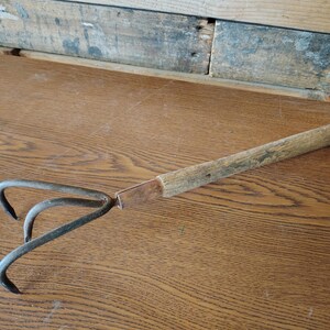 May include: A hand cultivator with three curved metal tines and a weathered wooden handle. The metal head is attached to the handle with a copper-colored metal ferrule. The tool features a rustic appearance with visible signs of age and use.