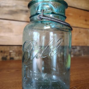 May include: A vintage aqua-colored glass jar with a wire bail closure. The front of the jar features the embossed text "Ball" in script and "IDEAL" in block letters, with additional small embossed text near the base of the cylindrical container.