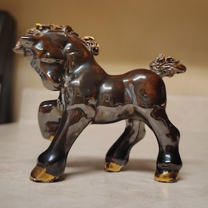 May include: A ceramic horse figurine in a standing pose. The horse has a glossy, dark brown body with gold accents on the hooves and mane. The figurine is on a light-colored surface.
