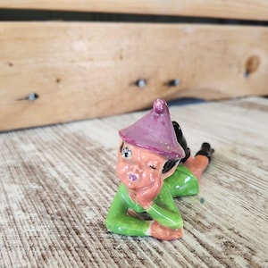 May include: A ceramic figurine of a reclining elf wearing a purple pointed hat and a green tunic. The elf has a peach-colored face, black hair, and is resting on one arm with a winking expression. The figure features glossy paint and black boots.