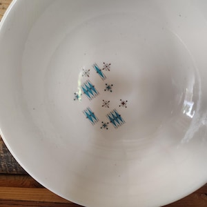 May include: A white ceramic bowl featuring a central pattern of small, repeating geometric shapes in bright blue and dark brown. The design consists of stylized diamond-like motifs and tiny cross-shaped accents arranged in a cluster at the base.