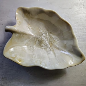 May include: A leaf-shaped ceramic dish with a glossy finish. The dish is primarily off-white with subtle brown and yellow accents, and a textured, crystallized pattern. The dish has a stem-like handle and is likely used for serving or display.