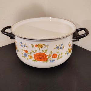 May include: White enamel cooking pot with black handles and a floral design in orange, yellow, blue, and green. The pot has a silver rim and sits on a dark surface. A vintage kitchenware item.