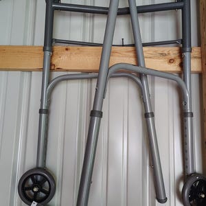 May include: A gray metal medical walker featuring two front wheels, two rear rubber-tipped legs, and black foam hand grips. The frame includes adjustable height settings on the legs and comes with an attached instructional manual and warranty booklet.
