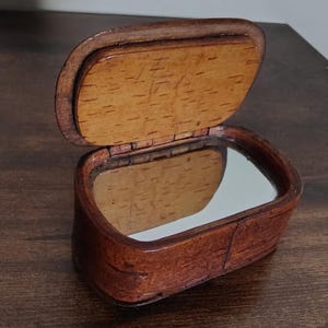 May include: A small, rectangular wooden box with a hinged lid, open to reveal a mirror. The box is made of dark brown wood with a lighter wood interior. The lid is slightly ajar, showing the mirror inside.