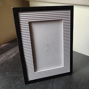 May include: A black and white photo frame with a textured, ribbed design. The frame has a rectangular opening for a photograph and is angled slightly. The frame is sitting on a dark surface.