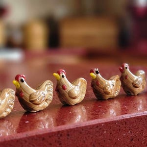 以下が含まれることがあります： 一列に並んだ6つの小さな陶器製の鶏の置物。それぞれの置物は、茶色の釉薬が塗られた体に、黄色い嘴、赤い鶏冠、黒い目という特徴があります。鶏は一列に配置されており、成形された羽の質感が細部まで表現されています。
