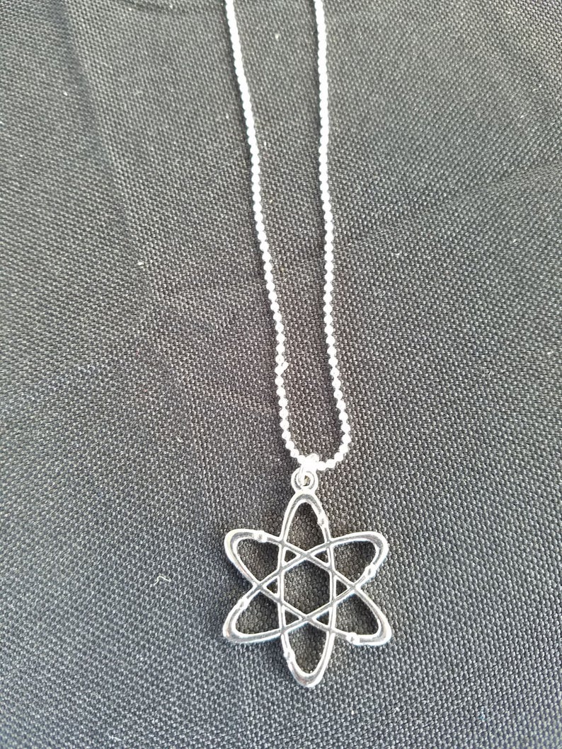 May include: A silver chain necklace with an atom pendant. The atom is a simple design with a circle in the center and lines extending out to form a star shape.