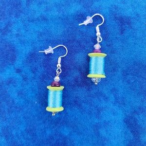 May include: A pair of earrings featuring a spooled thread design. The spools are made of light blue thread with a green base and a purple bead in the center. The earrings are silver with clear crystal accents.