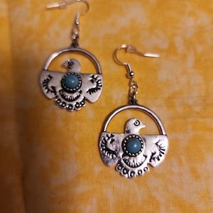 May include: A pair of silver earrings with a turquoise stone in the center of each earring. The earrings are shaped like a bird with its wings spread out.