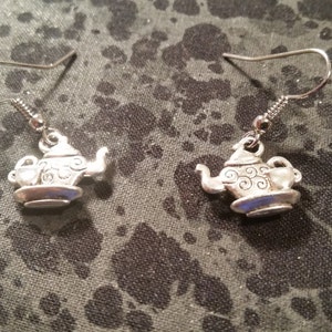 May include: A pair of silver teapot earrings with a small teacup on the side of the teapot. The earrings are on a silver hook.