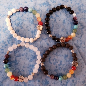 May include: Four bracelets with colorful gemstone beads and silver paw print charms. The bracelets are made with white lava rock, black lava rock, black onyx, and white jade beads.
