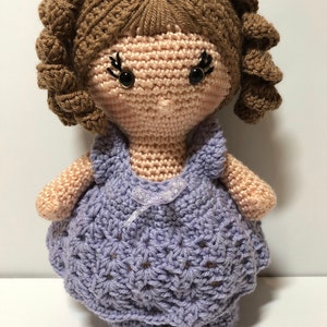 May include: A handmade crochet doll with brown curly hair and lavender accents. The doll has black button eyes and is wearing a lavender dress with a white ribbon. The doll is a soft, handcrafted item.