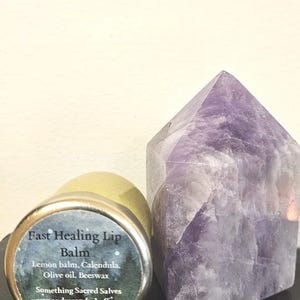 Può includere: Un barattolo di metallo rotondo di balsamo labiale Fast Healing con un'etichetta che elenca melissa, calendula, olio d'oliva e cera d'api, accanto a un grande cristallo di ametista viola a punta e un piccolo frammento di cristallo viola.