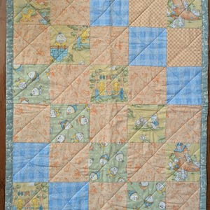 May include: A patchwork quilt featuring a grid of squares with illustrations of nursery rhyme characters, including Humpty Dumpty and rabbits, interspersed with light blue gingham and orange patterned fabric, finished with a sage green border.