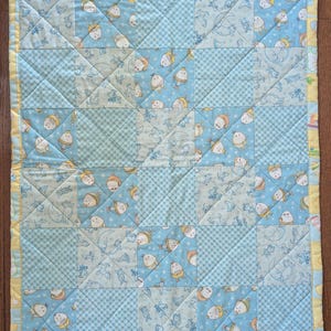 May include: A blue and yellow baby quilt with a patchwork design featuring a repeating pattern of little chicks wearing crowns. The quilt is quilted with a diamond pattern.
