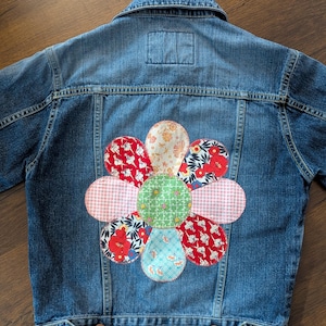 May include: A blue denim jacket featuring a large, multi-patterned fabric flower appliqué on the back. The flower consists of a green centre surrounded by eight petals in various colours and prints, including red, white, and blue floral and gingham designs.