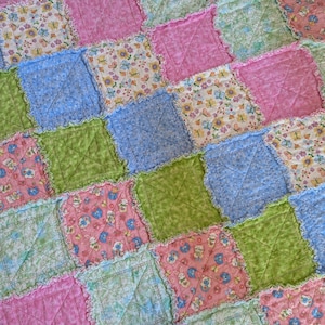 Peut inclure: Une courtepointe patchwork aux couleurs pastel. Elle présente des carrés roses, bleus et verts, avec des motifs de papillons et de lapins. Les bords de chaque carré sont froncés.