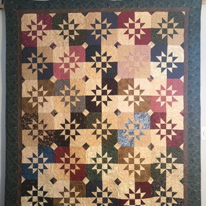 Peut inclure: Une courtepointe avec un motif de patchwork composé de tissus de différentes couleurs, notamment le brun, le vert, le rouge et la crème. La courtepointe est composée de carrés avec un motif d'étoile au centre de chaque carré.