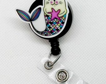 May include: A black retractable badge holder with a white cat mermaid design. The cat is holding a pink starfish and has a rainbow tail.