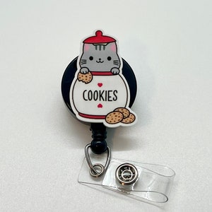 May include: A black retractable badge holder with a gray cat in a cookie jar design. The cat is holding a cookie and the jar says "COOKIES".
