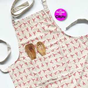 May include: A beige cotton apron with a red lobster print and a large pocket. The apron has adjustable straps and is ideal for cooking or baking.