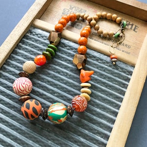 May include: A colorful necklace with orange, brown, white, and green beads. The necklace is made of wood, metal, and ceramic beads. The necklace is on a wooden surface with the text "Do Not Rub Hard" printed on it.