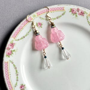 May include: A pair of pink gemstone earrings with gold-colored hooks. Each earring features a carved pink Buddha figure, a small gold bead, and a clear, bell-shaped crystal. The earrings are displayed on a floral-patterned plate.