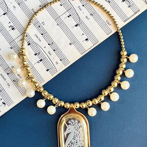 May include: A gold necklace with a unicorn pendant. The necklace is made of gold beads and has white pearl accents. The pendant is oval shaped and features a white unicorn with a floral design.