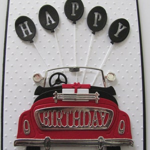 May include: A red vintage car with a silver "Birthday" number plate. Five black balloons with white letters spelling "Happy" are attached to the car.