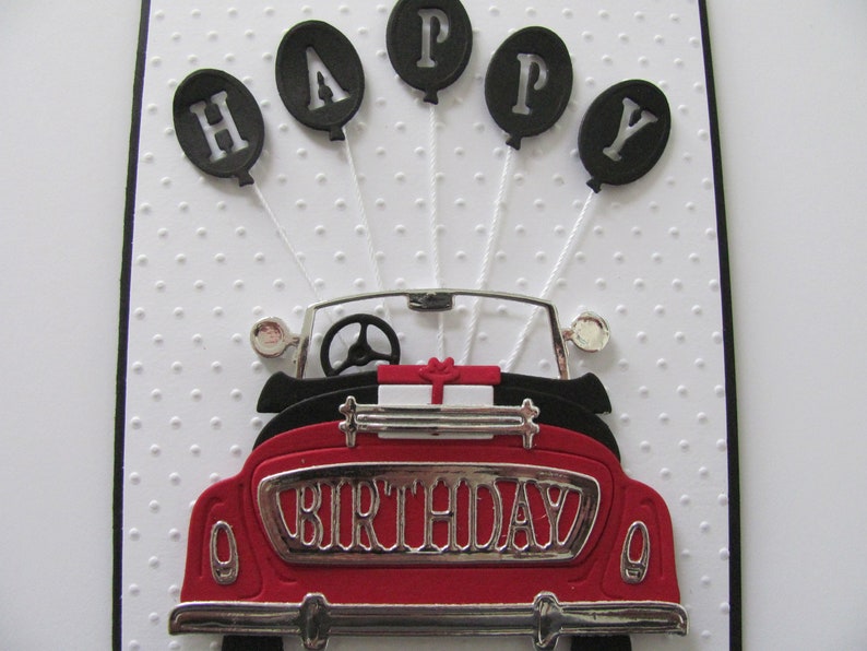 May include: A red vintage car with a silver "Birthday" number plate. Five black balloons with white letters spelling "Happy" are attached to the car.