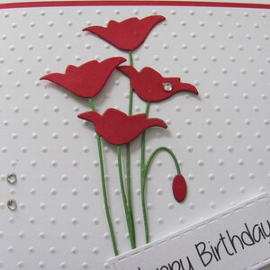 May include: A white card with a red border and a red dotted pattern. The card has a red paper flower bouquet with green stems and a white label with the text "Happy Birthday".