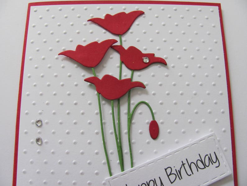 May include: A white card with a red border and a red dotted pattern. The card has a red paper flower bouquet with green stems and a white label with the text "Happy Birthday".