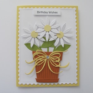 May include: A white card with a yellow border and a scalloped edge. The card has a white background with a yellow banner at the top that says "Birthday Wishes". The card features a paper craft design of three white daisies in a brown pot with a yellow bow.