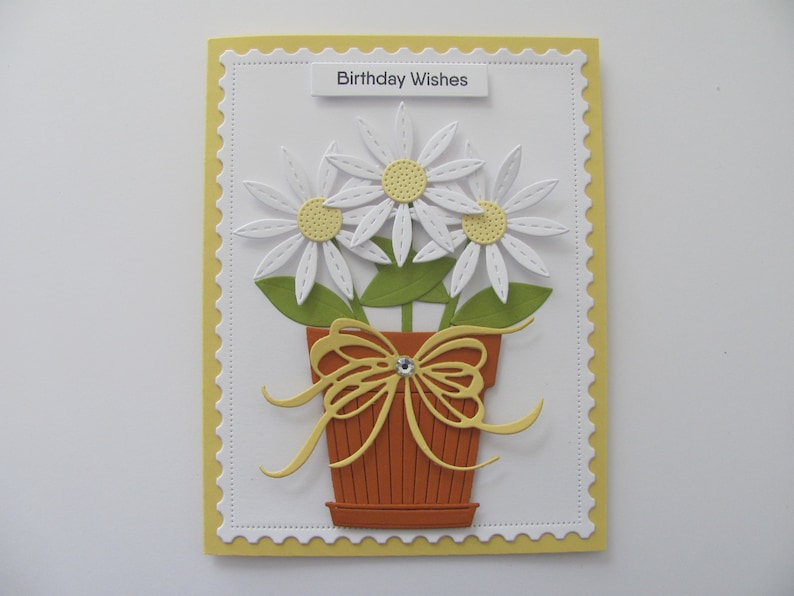 May include: A white card with a yellow border and a scalloped edge. The card has a white background with a yellow banner at the top that says "Birthday Wishes". The card features a paper craft design of three white daisies in a brown pot with a yellow bow.