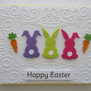 May include: A white card with a yellow border and a white embossed design. Three paper bunnies in purple, green, and pink are arranged in a row with a carrot on each side. The card reads "Happy Easter".